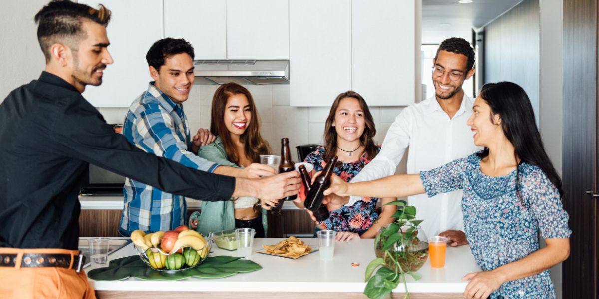Group of friends smiling and clinking drinks together in a kitchen during a housewarming party.