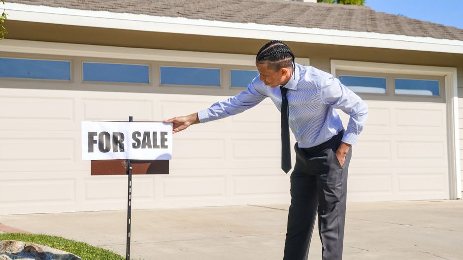A professional 'For Sale' sign is posted in the manicured front lawn of a charming suburban home on a sunny day.